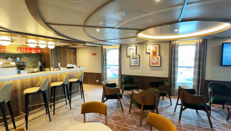 The 'living room' style area aboard the Seabourn Venture, designed to provide a cozy and inviting ambiance. The space is elegantly appointed with a variety of seating options, including high stools at the cafe, plush green and mustard armchairs, and wooden chairs grouped around small tables. The area is highlighted by soft, circular lighting overhead and stylish decor elements such as modern art pieces framed on the walls. This setup creates a relaxed, social atmosphere where guests can enjoy drinks, converse, or simply unwind during their voyage.