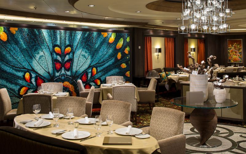 The upscale 150 Central Park restaurant on Harmony of the Seas cruise ship features elegant dining arrangements with beige upholstered chairs and crisp white tablecloths. The room is warmly lit, highlighted by an eye-catching, vibrant butterfly wing mosaic art on the wall, providing a luxurious dining atmosphere.