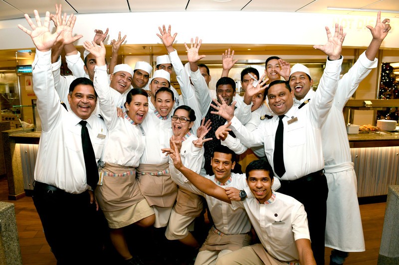 A group of cheerful cruise ship staff poses together, raising their hands and smiling widely. They are dressed in uniforms, with some in white shirts and black ties, and others in kitchen attire, standing in what appears to be a dining or buffet area.