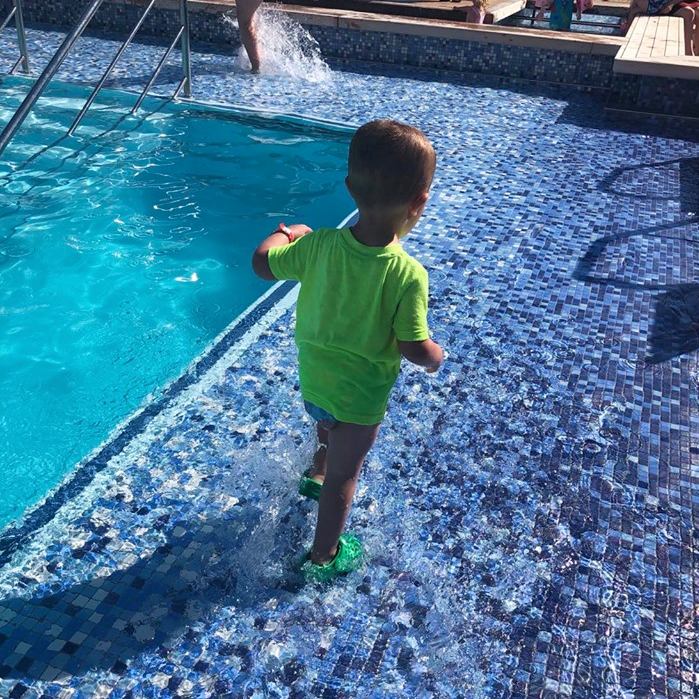 A small child in a bright green shirt and swim nappy cautiously steps into the sparkling blue mosaic-tiled pool on a cruise ship, representing the careful curiosity of young swimmers.