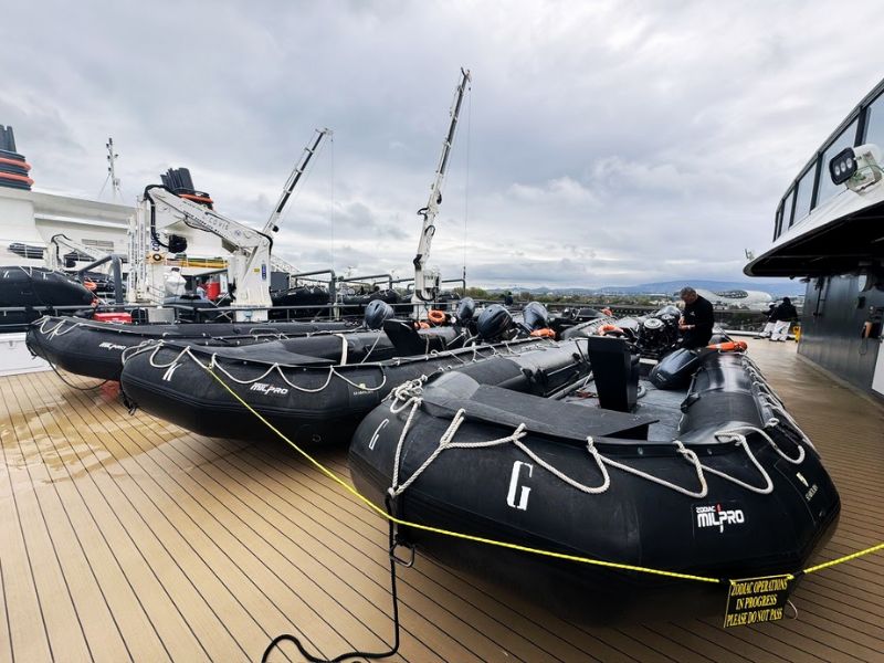 On the deck of the Seabourn Venture, several large black Zodiac boats are lined up, each secured and ready for deployment. These boats, essential for shore excursions and close-up wildlife viewing, are arranged neatly against the backdrop of a cloudy sky. A crew member is seen working on one of the boats, emphasizing the ongoing preparations for guest activities.