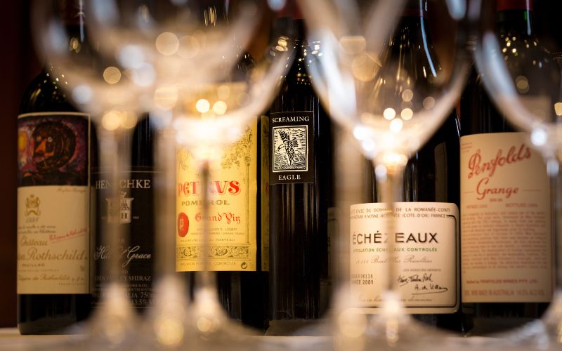A selective focus shot of various high-end wine bottles, including labels such as Screaming Eagle and Penfolds Grange, viewed through the rim of a wine glass. The setting suggests a luxurious assortment, possibly on a Cunard cruise ship.