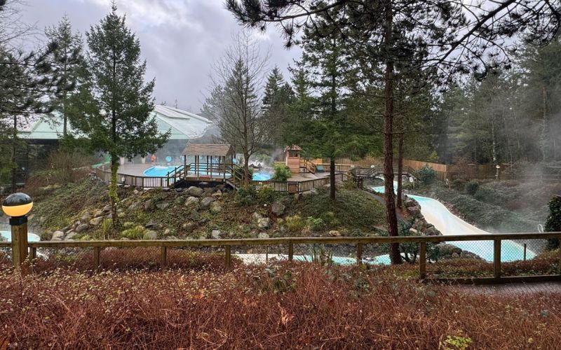 A view of the outdoor pool complex at Center Parcs surrounded by lush evergreen trees and shrubs with a hint of steam rising from the warm water on a chilly day, showcasing the resort's integration with nature and providing an inviting atmosphere for relaxation and recreation.