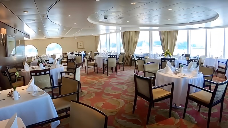 This image depicts an elegantly set cruise ship dining room with a panoramic view from large windows. The room features neatly arranged tables with white linen, surrounded by comfortable chairs, and complemented by a stylishly patterned carpet. The overall ambiance is inviting, with a sense of spaciousness and luxury, ideal for a formal dining experience at sea.