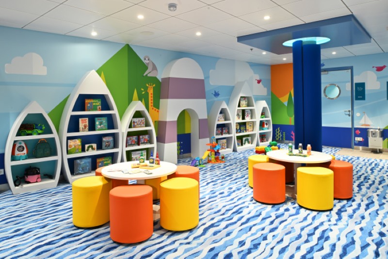 The colorful and inviting Adventure Ocean kids' area on Icon of the Seas, designed with boat-shaped bookshelves, bright geometric shapes, and playful seating on a wavy blue carpet, creating a fun learning and play environment for children.