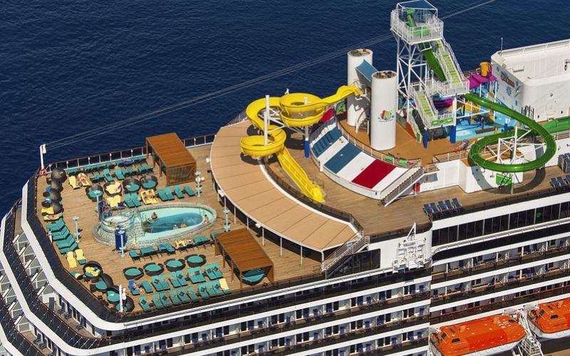 Aerial view of the vibrant Aft Serenity Deck on the Carnival Pride, showcasing a bustling pool area, colorful water slides, an adventure ropes course, and ample lounging options with the deep blue ocean stretching into the horizon.