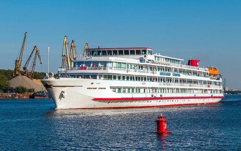 The river cruise ship 'Aleksandr Suvorov' glides on calm waters, with its white and red exterior and multiple decks visible against a backdrop of industrial cranes and a clear blue sky.