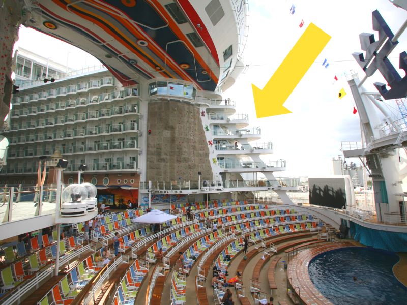 View of Allure of the Seas' AquaTheater Suites, indicated by a yellow arrow, with a bustling amphitheater seating area and a large outdoor screen overlooking the ship's wake.