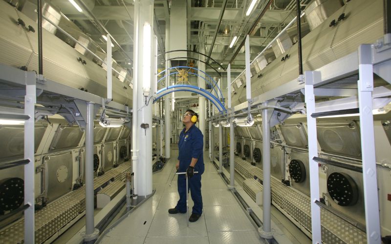 An engineer in blue coveralls and ear protection standing inside the engine room of Royal Caribbean's Allure of the Seas, surrounded by machinery.