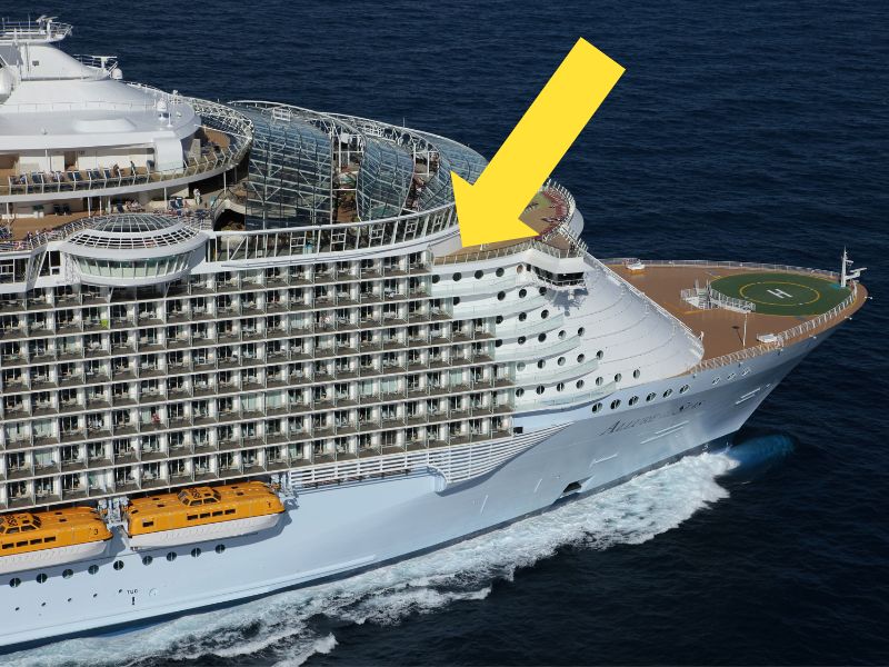 Aerial view of Allure of the Seas cruise ship showcasing the starboard side, with a yellow arrow pointing to balcony rooms potentially prone to seasickness and wind exposure, while cruising on open sea.