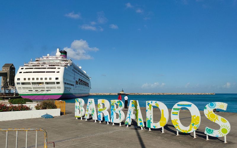 The Ambience cruise ship is docked at a port in Barbados, under a clear blue sky. The foreground features large, colorful letters spelling "BARBADOS," decorated with tropical designs. The scene captures the vibrant and welcoming atmosphere of the Caribbean island.