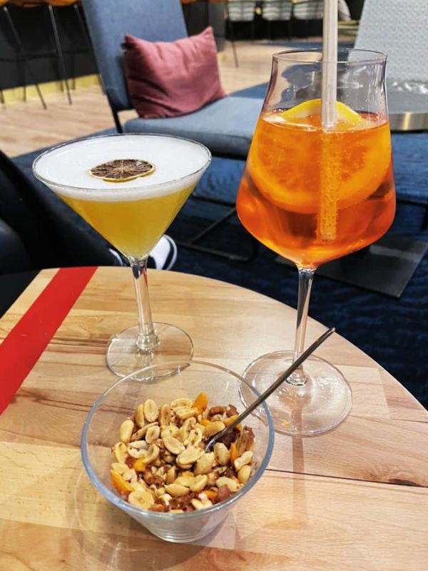Two cocktails on a wooden table aboard a cruise ship: a yellow drink with a dried citrus garnish and an Aperol Spritz with an orange slice. A bowl of mixed nuts is also on the table, with cozy seating and decorative pillows in the background, creating a relaxed and inviting atmosphere.