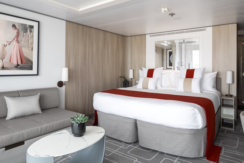 AquaClass Sky Suite on a Celebrity cruise ship, featuring a serene and stylish decor with a plush bed, accented with red and beige, a comfortable sofa, and a framed picture of an elegant vintage dress, enhancing the luxurious cabin experience.