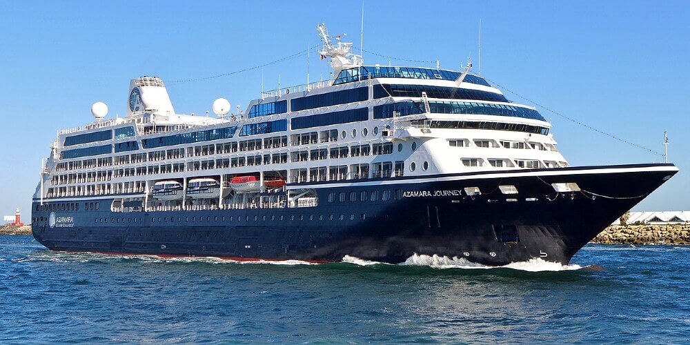 The Azamara Journey cruise ship captured in full motion, cutting through the waters with its distinctive dark hull and white superstructure. The ship is elegantly designed with multiple decks visible, lined with balconies and windows, under a clear blue sky. This vessel is shown near a breakwater, indicating its approach to or departure from a port, with its wake trailing behind, emphasizing its forward movement and dynamic presence in a vibrant maritime setting.