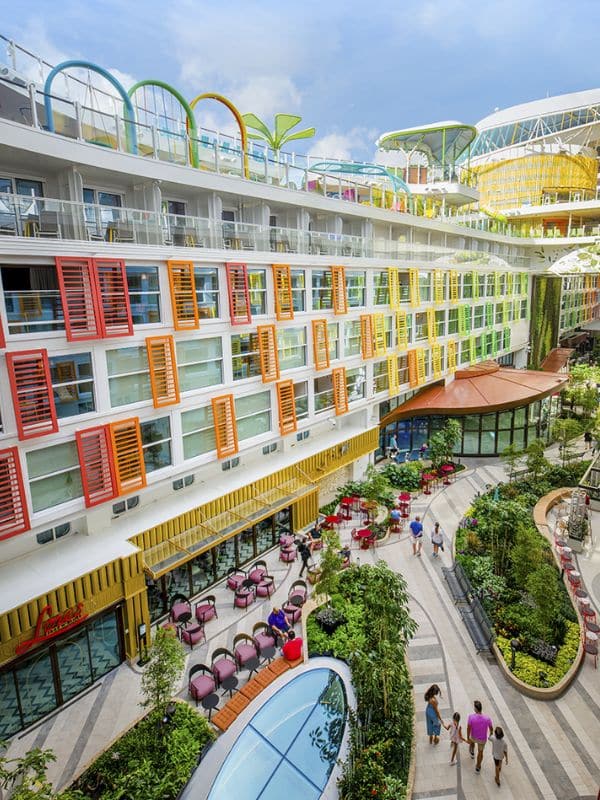 Lively central park area inside Royal Caribbean's 'Icon of the Seas,' featuring multiple levels of staterooms with colorful shutters in red, orange, and yellow. The open-air atrium is bustling with activity, adorned with green plants, outdoor seating areas, and a curving pathway, creating a vibrant community space onboard the cruise ship.
