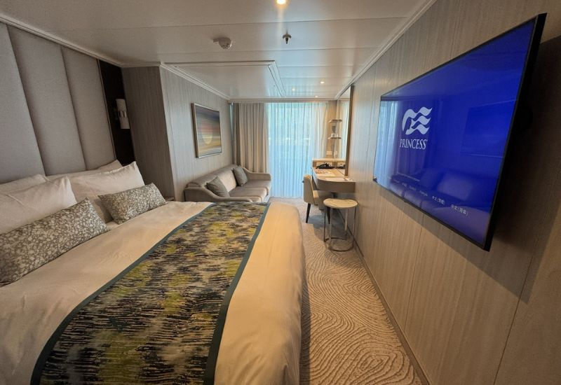 The image shows a Balcony Stateroom on Princess Cruises' Sun Princess. The room features a large bed with white linens, decorative pillows, and a patterned bed runner. A wall-mounted flat-screen TV displays the Princess Cruises logo. The stateroom includes a sofa, a desk with a chair, and a small table, all arranged near a sliding glass door that opens onto the balcony. The decor is modern and stylish, with neutral tones and soft lighting that creates a relaxing atmosphere, perfect for enjoying the ocean views from the balcony.