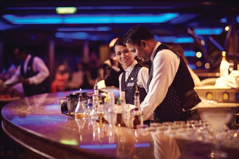Two bartenders are working behind a well-stocked bar on a cruise ship. They are dressed in matching uniforms, with one bartender preparing a drink while the other smiles and looks on. The bar area is illuminated with ambient blue lighting, and various bottles and mixers are neatly arranged on the counter, creating a vibrant and inviting atmosphere for the guests.