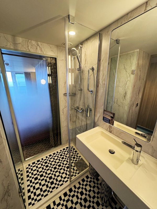 Modern bathroom interior on the Costa Smeralda cruise ship's balcony room, featuring a glass-enclosed shower, marble walls, and a black-and-white checkered floor.