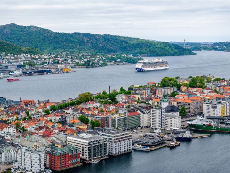 The 'Sky Princess' cruise ship from Princess Cruises is seen in the distance, set against the backdrop of a coastal cityscape with colorful buildings and rolling hills, likely a port of call on one of its voyages.