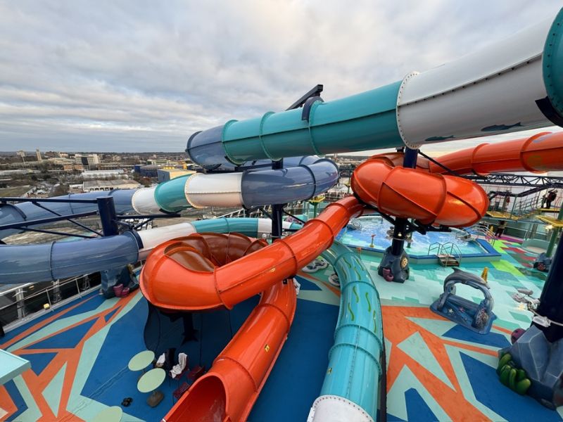 MSC Euribia aft deck featuring a colorful array of winding waterslides in blue, teal, and orange. The slides spiral above a playful water park area, with a cityscape visible in the distance under a cloudy sky.