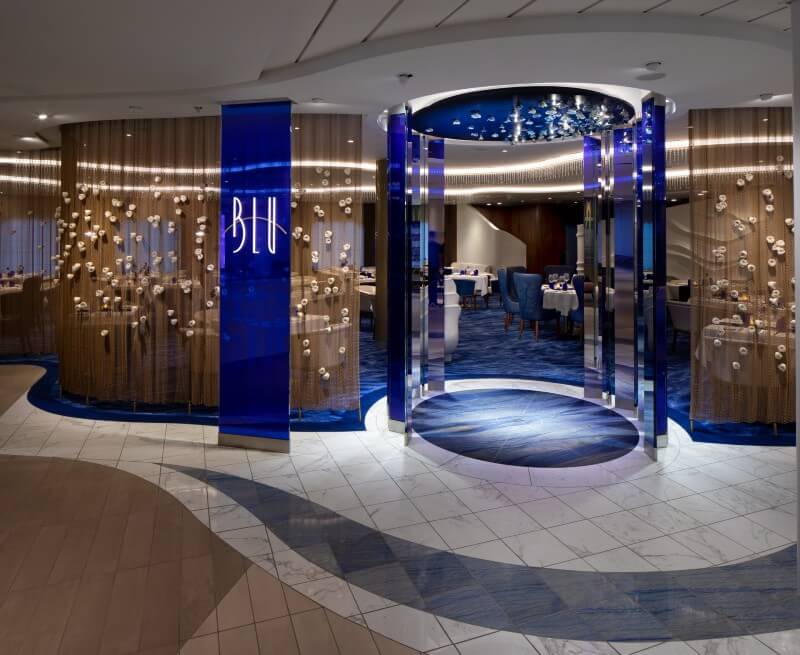 Entrance to Blu restaurant on a Celebrity cruise ship, featuring elegant modern decor with a circular blue light installation, 'Blu' signage in bright neon, and a stylish dining area with chic furnishings that evoke an upscale, serene oceanic atmosphere.