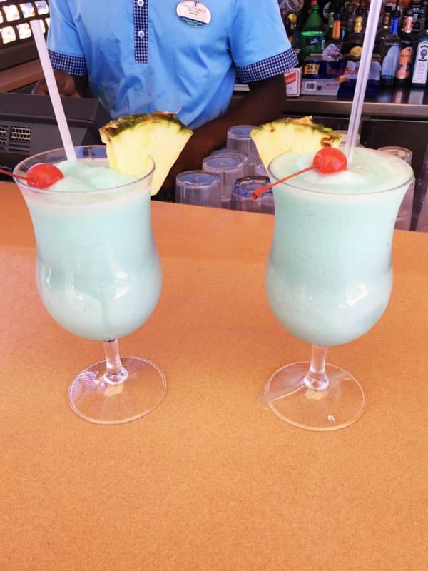 Two Blue Hawaiian cocktails on a cruise ship, served in hurricane glasses and garnished with pineapple slices and cherries. The drinks are placed on a bar counter, with a bartender in a blue uniform in the background, and various bottles and bar equipment visible, creating a lively and tropical bar atmosphere.