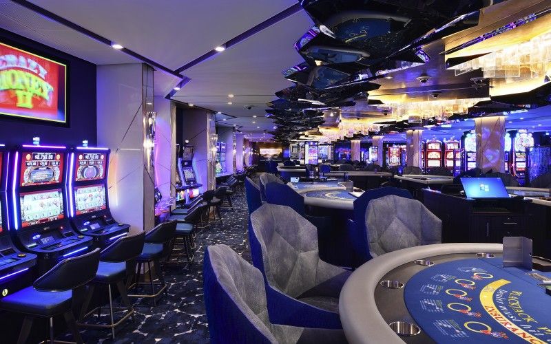 A sophisticated casino setting showing an array of slot machines on one side and card game tables on the other, all under elegant, modern lighting. The plush blue chairs and the dark carpet add to the luxurious ambiance of the gambling floor.