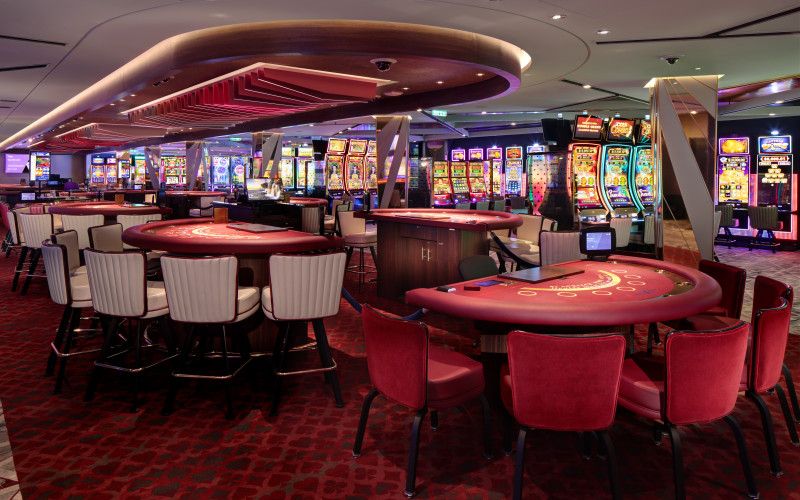 A Celebrity casino floor elegantly designed with red and white gaming tables in the foreground and a selection of slot machines in the background. The ambient lighting and vibrant carpet pattern create an inviting atmosphere for gaming enthusiasts.