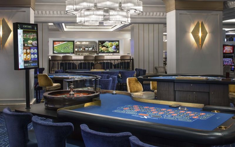 A refined casino interior with a bar in the background, gaming tables in the center, and comfortable blue chairs around, illuminated by a grand chandelier. The scene is set for an evening of sophisticated gaming and socializing.
