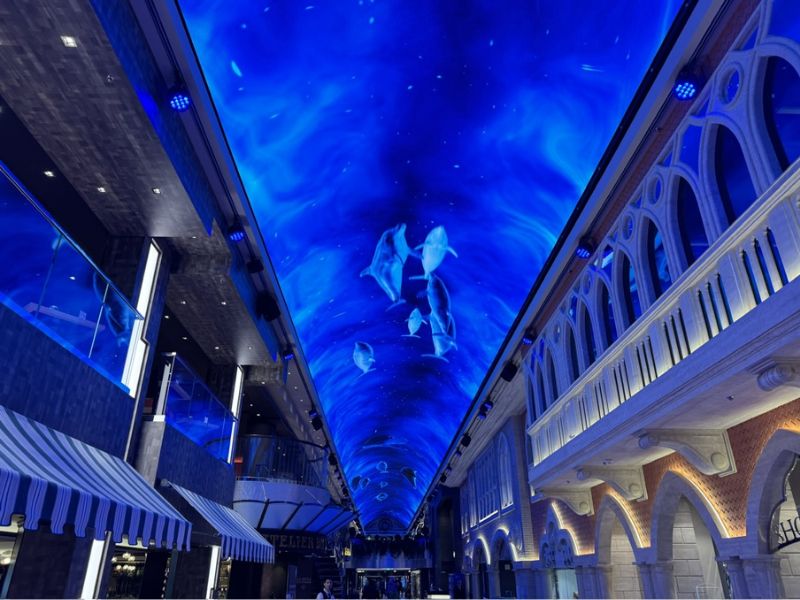 An enchanting cruise ship promenade on MSC Euribia illuminated by a ceiling LED screen displaying a vibrant underwater scene with swimming stingrays. The area is architecturally detailed with arched walkways and shopfronts, creating an immersive, oceanic shopping experience.