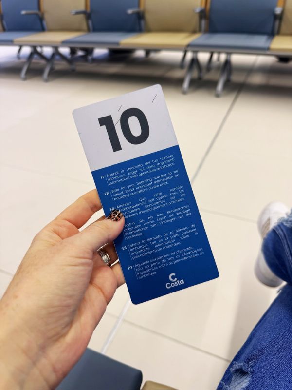 Cruise Mummy holding a blue Costa boarding card with the number 10, in a waiting area with seats in the background, indicating the boarding process on the Costa Smeralda cruise ship. The card includes multi-language instructions for boarding.