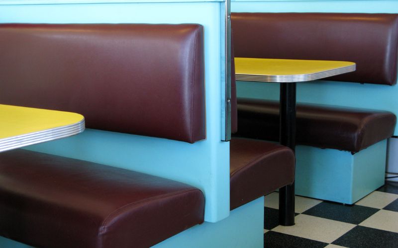 This image showcases booth-style seating typical of a retro diner, with maroon and blue vinyl benches and yellow tabletops. The background features a classic black and white checkered floor, enhancing the nostalgic feel of the setting. This arrangement is characteristic of mid-20th century American diners, offering a cozy and casual dining experience.