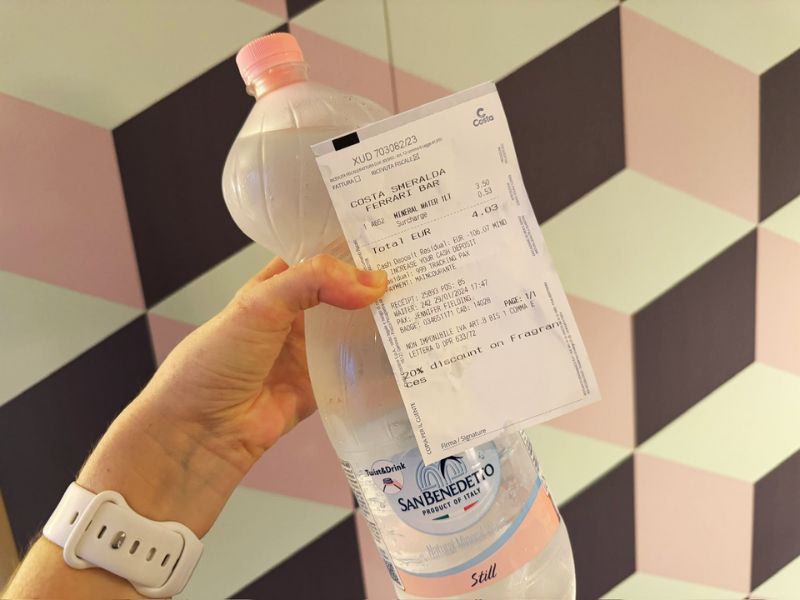 Cruise Mummy holds a transparent bottle of San Benedetto still water with a Costa Smeralda receipt attached, indicating a recent purchase onboard the cruise, set against a geometrically patterned pastel background.