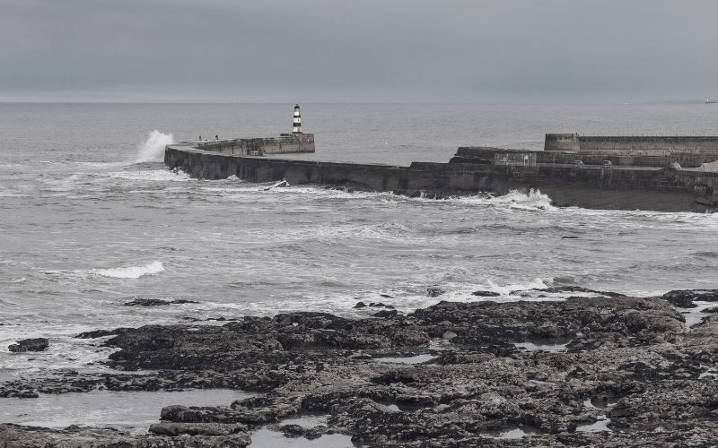 The image shows a rugged British coastline under a cloudy sky, with a stone pier extending into the choppy sea. A striped lighthouse stands at the end of the pier, and waves crash against the structure, creating a moody and atmospheric scene. The foreground features dark, rocky terrain, contrasting with the gray sea and sky.