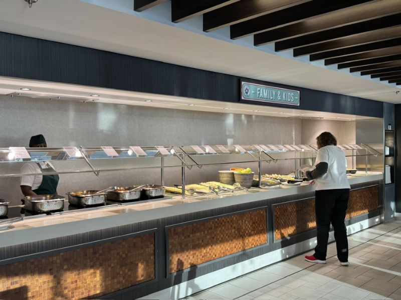 A guest at a cruise ship buffet under the 'Family & Kids' sign, serving themselves from a variety of dishes. The buffet area is clean and well-organized, featuring a selection of fruits and warm foods, with a crew member in attendance.