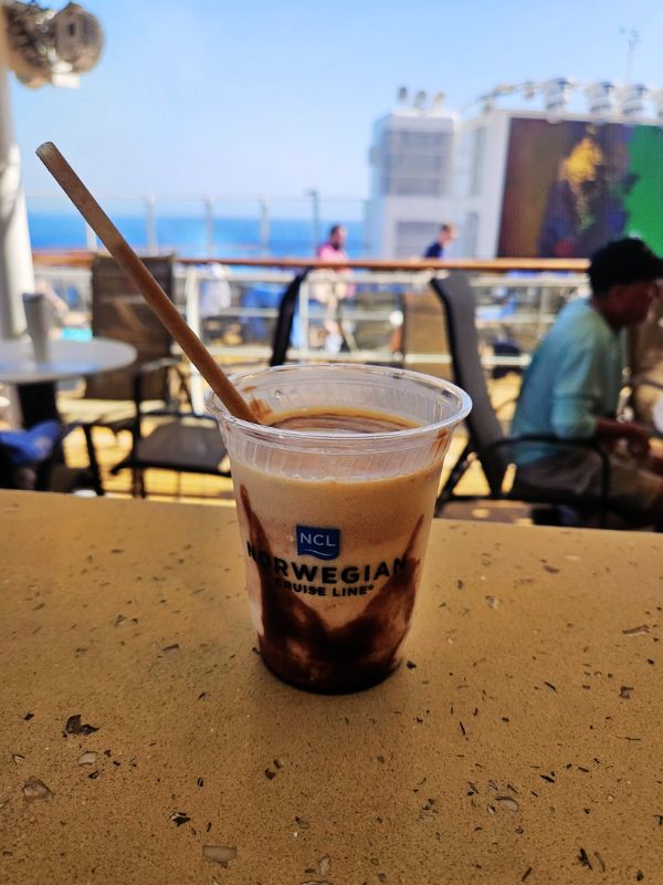 A Bushwacker cocktail in a clear plastic cup with the Norwegian Cruise Line logo, featuring a swirl of chocolate syrup. The drink is set on a table with a pool deck in the background, capturing a sunny day at sea with passengers relaxing and enjoying the cruise amenities.