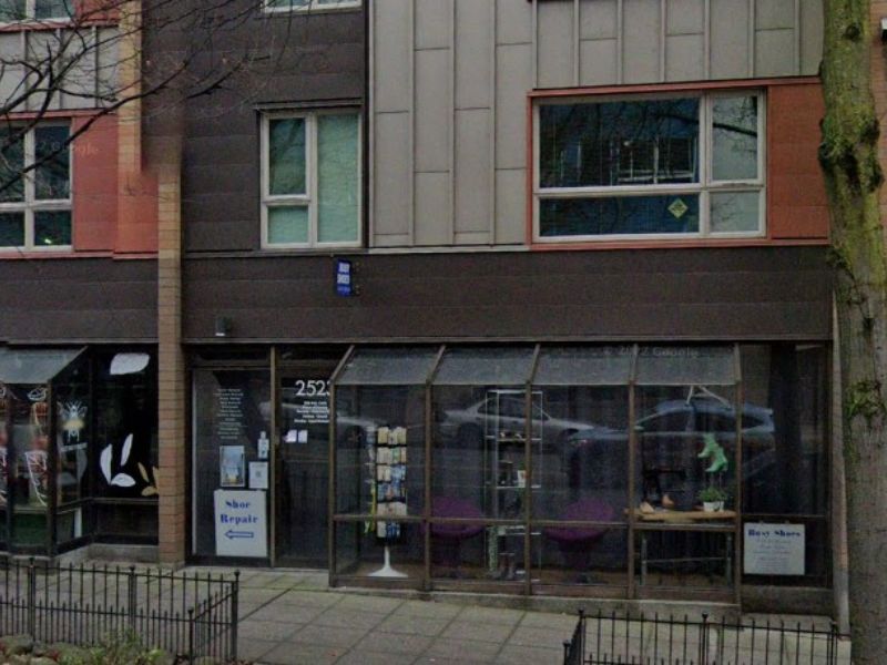 A shoe repair shop with a visible sign reading "Shoe Repair" is nestled between buildings, showcasing a variety of shoes in its window display on a cloudy day, suggesting a local business offering footwear services.