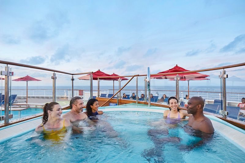 Guests enjoying a hot tub on the deck of a Carnival cruise ship, with ocean views and umbrellas in the background.