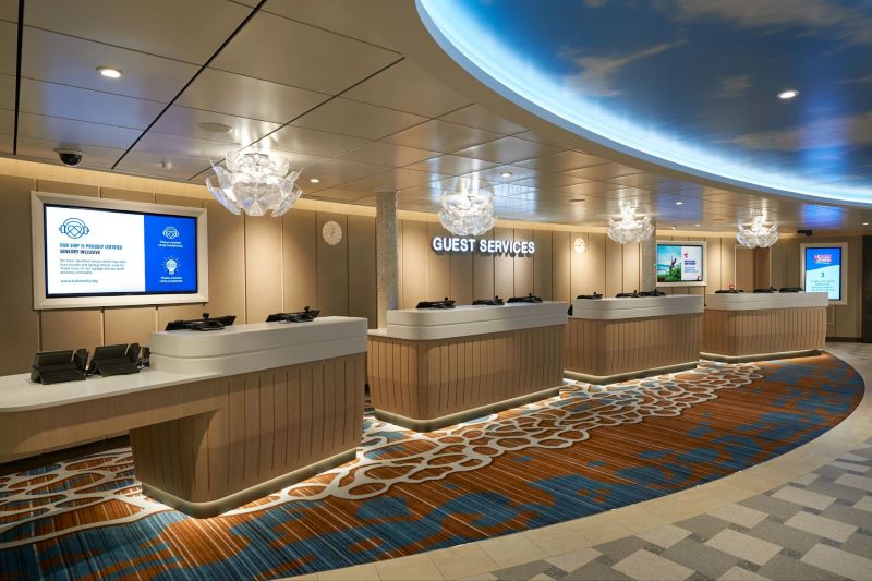 The modern and welcoming Guest Services desk on a Carnival Cruise ship, featuring a curved counter with several computer terminals, informational screens, and elegant lighting, ready to assist passengers with their inquiries.