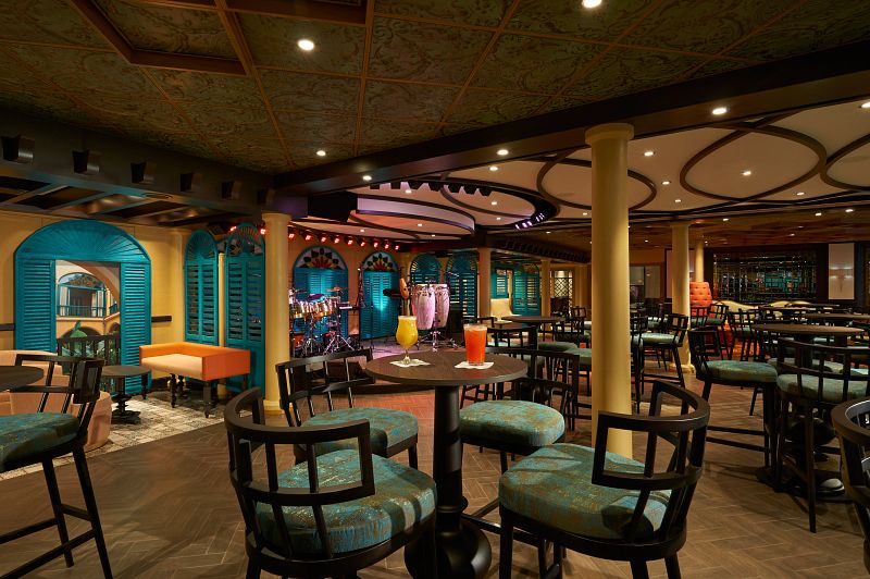 Warm and inviting Havana Bar on the Carnival Jubilee, featuring teal window shutters, ornate ceiling patterns, high stools at the bar tables, and refreshing tropical drinks, all contributing to a relaxed Cuban-inspired ambiance.