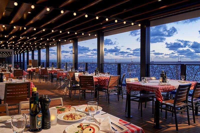 Evening ambiance at Cucina del Capitano on Carnival Mardi Gras, featuring an outdoor dining area with red checkered tablecloths, wooden chairs, and a serene ocean view framed by large windows under a dusky sky.