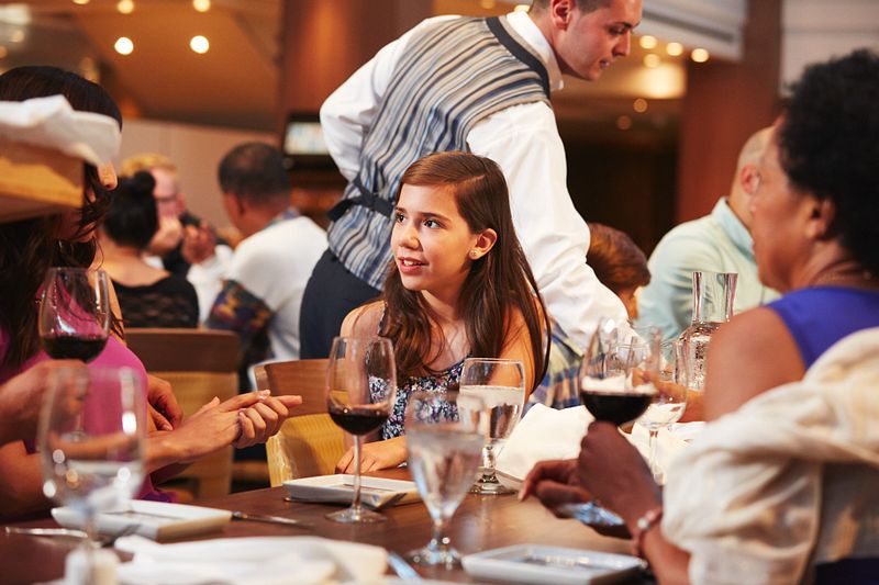 A young girl engaging in conversation at a family dinner in the main dining room on Carnival Vista, with waitstaff attending to guests and wine glasses on the table, capturing a moment of elegance.