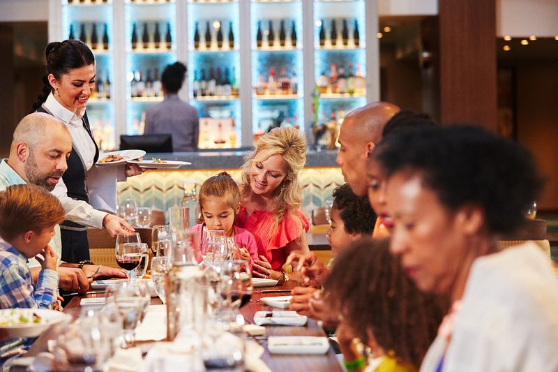 A vibrant scene in the main dining room on Carnival Vista, where a waitress is serving a family. The warm lighting and the restaurant's modern decor create a welcoming atmosphere for guests enjoying their meal.