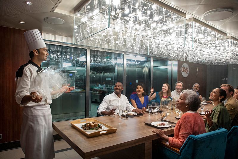 Guests at the Chef's Table on a Carnival cruise ship are treated to an engaging culinary demonstration by a smiling chef, set in an intimate dining space with modern glass and crystal décor, highlighting an exclusive and interactive gourmet experience.