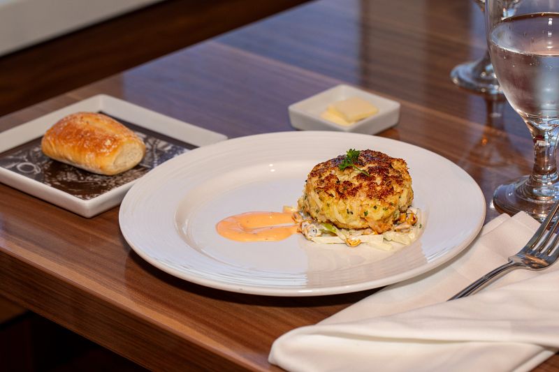 An elegantly plated crab cake served on a white plate with a dollop of sauce, accompanied by a freshly baked roll on a side plate and a glass of water, ready for a fine dining experience on a Carnival cruise ship's main dining room.