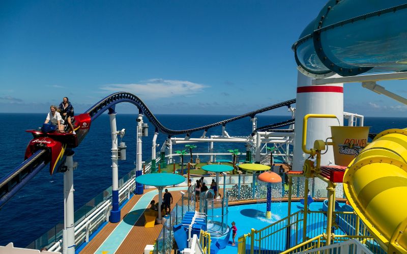 The Mardi Gras cruise ship's top deck is bustling with excitement, featuring the first roller coaster at sea, 'BOLT.' Passengers are enjoying a thrilling ride in a red car on the blue track, with the expansive blue ocean in the background. The deck also hosts a colorful water park area, with guests enjoying the sun and fun activities.