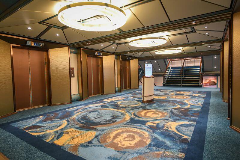 An elegant elevator area on the Carnival Radiance with a patterned blue carpet, wood-paneled doors, and modern circular light fixtures creating a welcoming atmosphere for guests transitioning between the cruise ship's multiple levels.