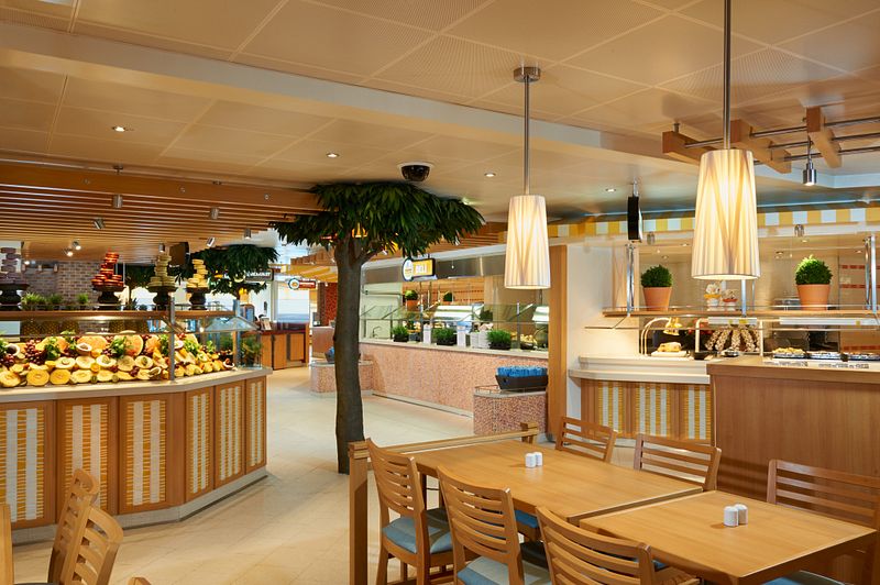 The Lido Marketplace on Carnival Vista, presenting a bright and airy buffet area with wooden tables, an array of fresh fruits on display, and elegant pendant lighting, offering a casual dining atmosphere.