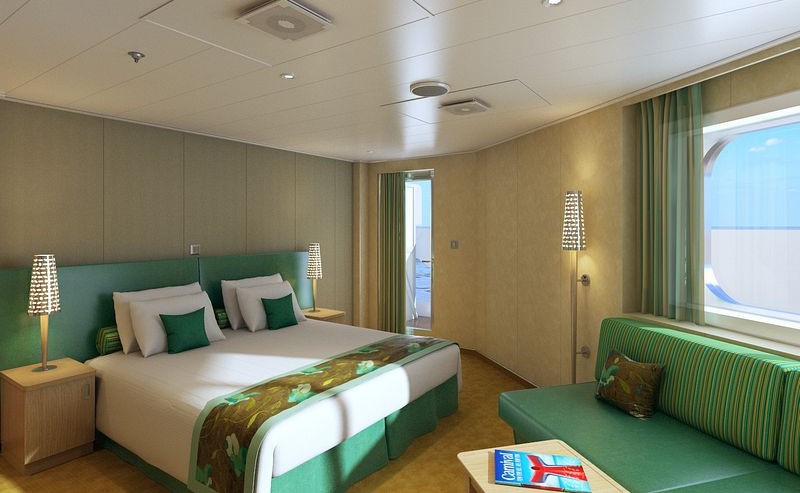 Cloud 9 Suite on Carnival Vista, showcasing a large bed with green accents, matching sofa, elegant wall-mounted lights, and a window offering natural light and sea views, creating a serene and comfortable space for guests.