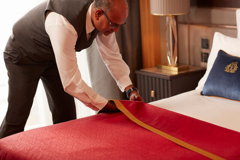 A diligent butler on a Cunard cruise ship meticulously prepares a stateroom, showcasing his attention to detail while arranging a red bed runner on crisp white bedding, embodying the line's commitment to luxury and service.